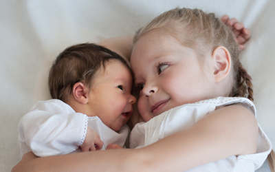 A young girl gently cuddles a baby. They are lying face to face on a bed