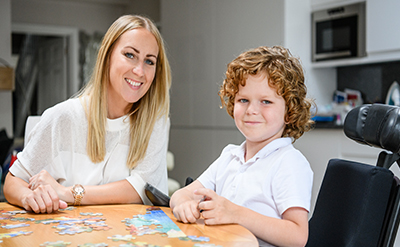A blonde boy and his foster carer smile into the camera. They are doing a jigsaw puzzle together