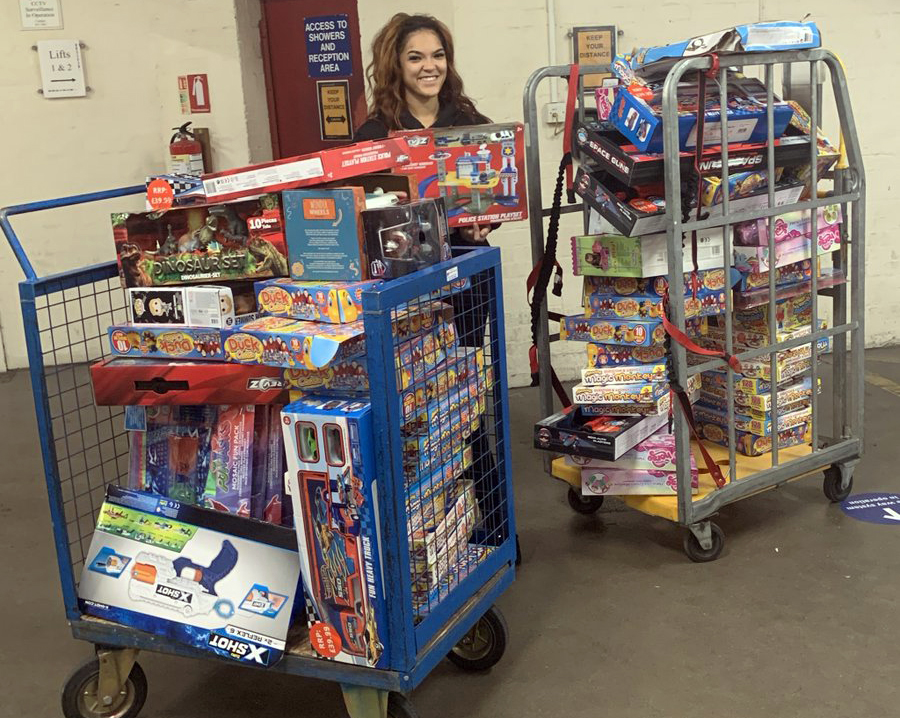 A staff member at Birmingham Children's Trust smiles at the camera, she is standing between two large retail trolleys filled with presents which are being delivered to the Trust's Winter Hub