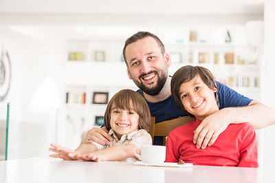An adoptive dad has his arm round with his two children. they are sitting at the kitchen table and smiling at the camera
