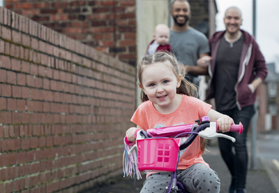 A girl rides her pink bike towards the camera. Her two dads walk behind her, carrying her baby brother