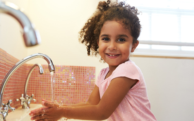 A girl washes her hands in the sink. she is wearing a pink top and had thick, curly hair