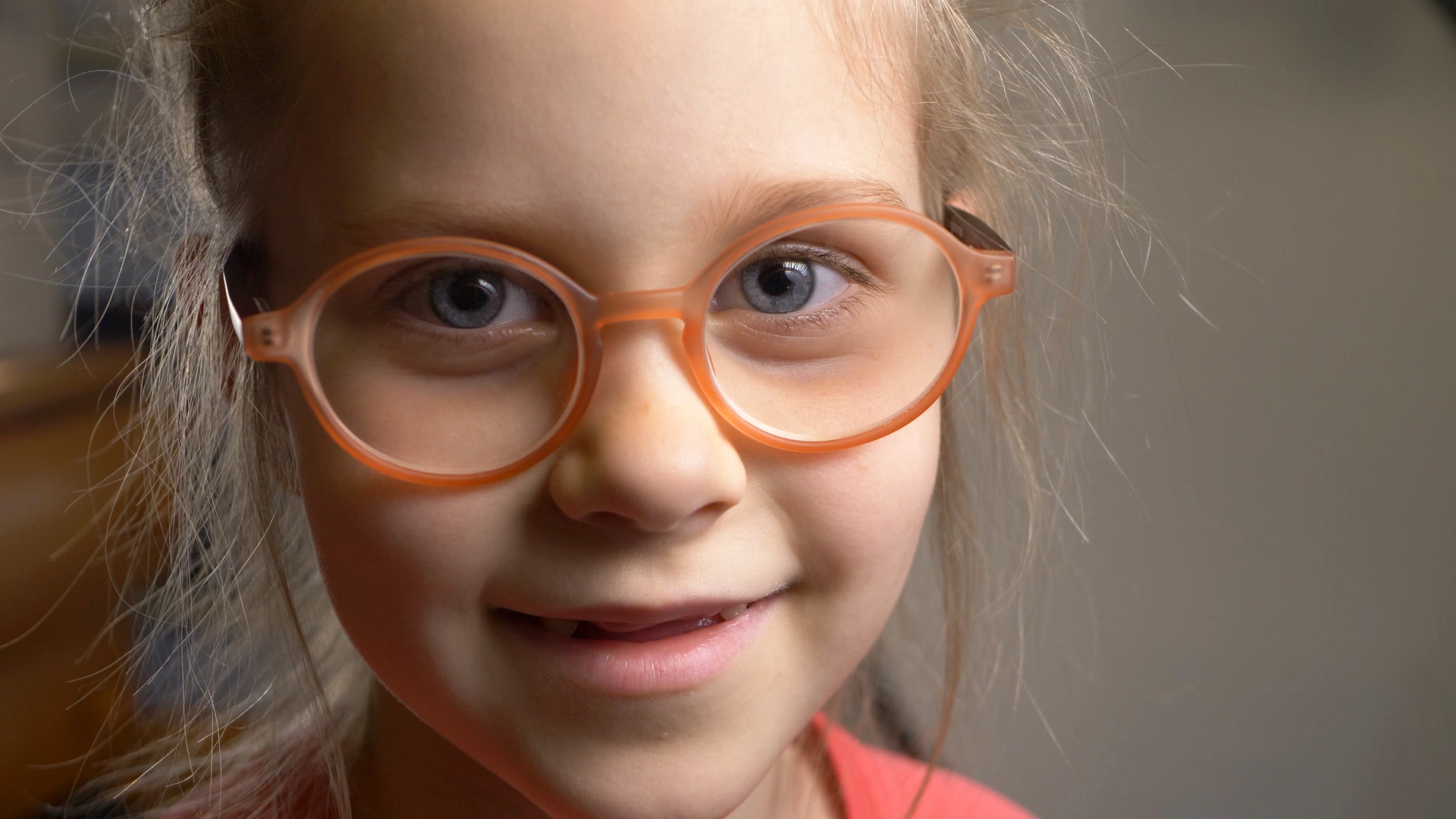 A young girl smiles into the camera, she is wearing round orange glasses and has a gappy smile where she has lost her milk teeth