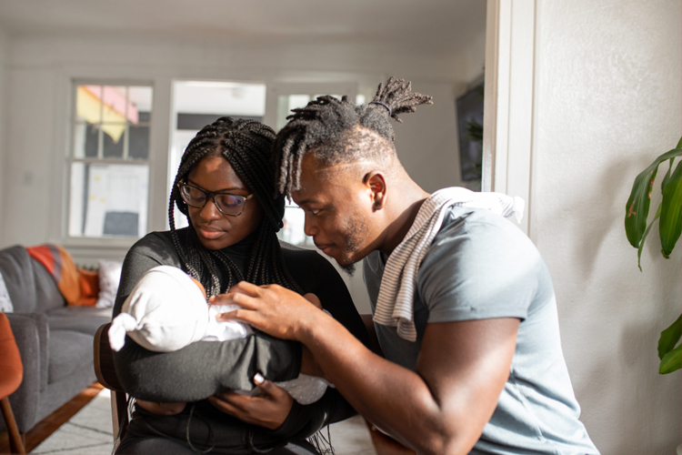 Two foster carers hold the young baby in their care. The female carer has long braids and is holding the baby. The male carer has a high top hairstyle and is leaning forwards to look at the baby's face