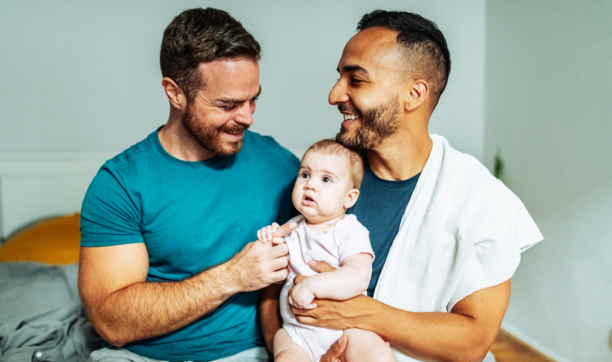Two dad are sitting on the edge of a bed, they are holding their baby between them and smiling