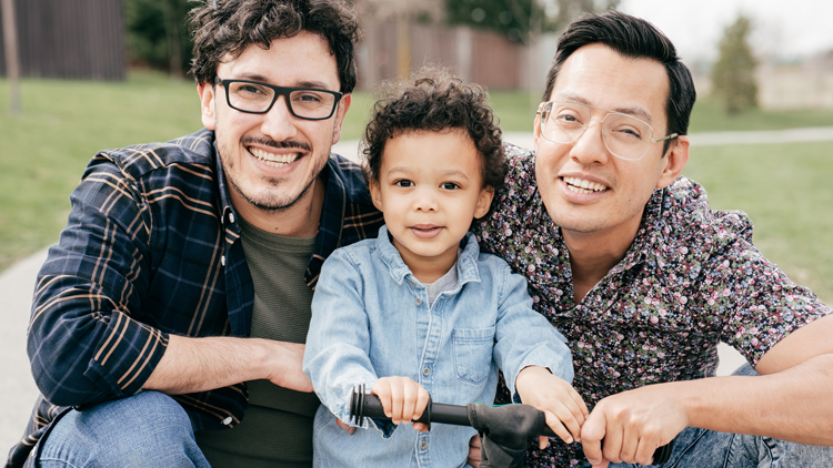 Two dads crouch down and smile at the camera, their young son stands between them with his push bike.