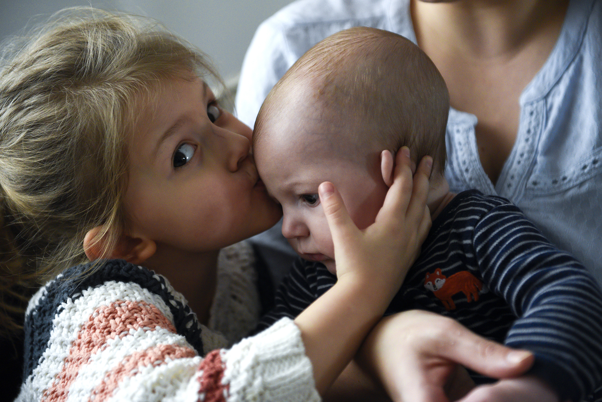 A young girl child kisses her baby brother's head. The baby is leaning towards his sister and is propped up on his mother's lap