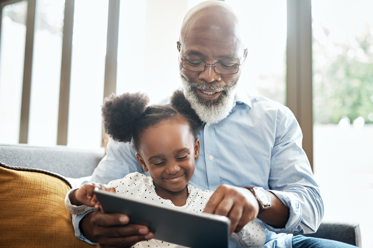 An older gentleman with a white beard, is sitting on the sofa with his Great Niece. He is supervising her whilst she plays on her tablet.  She is smiling and wearing her hair in space buns
