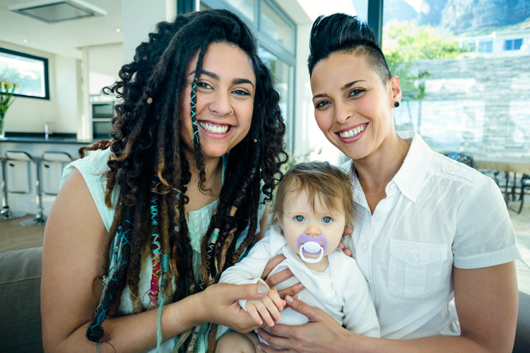 Two women smile into the camera. They are holding their baby daughter between them, she has a dummy in her mouth.