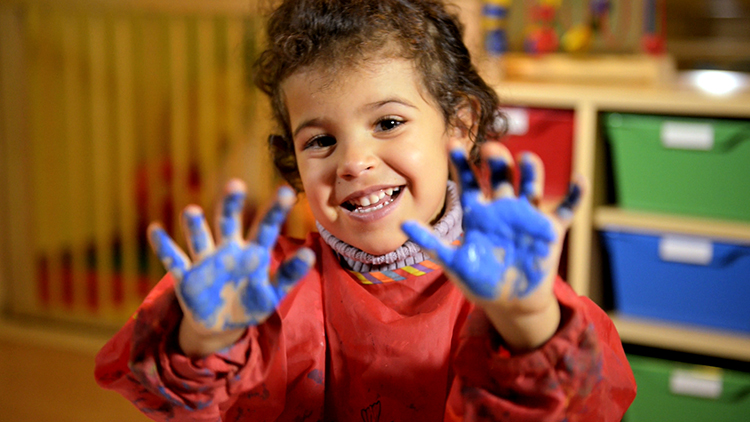 A four year old girl smiles into the camera. She is holding up her hands which are covered in blue paint.
