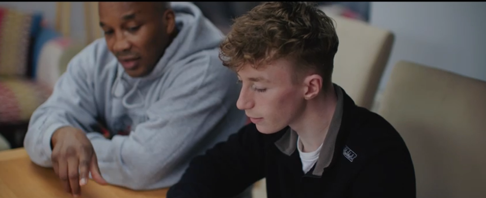 A foster carer and the young person he looks at are sitting at the kitchen table. The foster carer is pointing to the boy's homework and helping him through it.