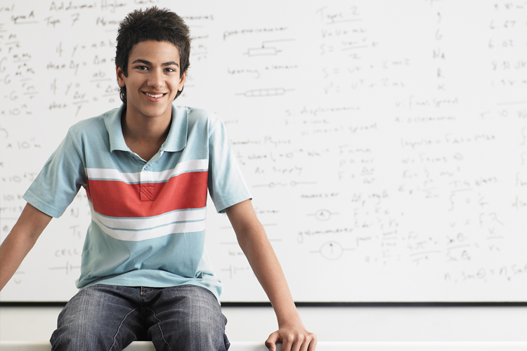 A teenage boy sits in front of a white board covered in equations. He is smiling into the camera
