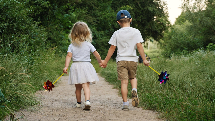 An older brother and his little sister are holding hands. They are walking down a sandy path with their backs to the camera.